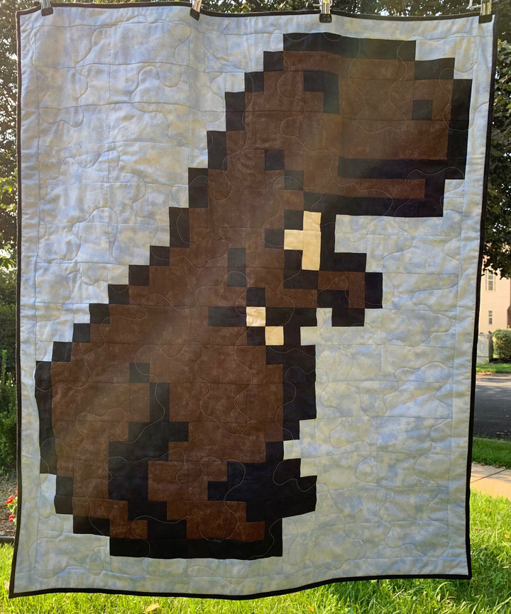 Finished T-Rex pixel quilt in brown and tan dinosaur fabrics on a cloud-print sky background, machine quilted, photographed hanging outdoors